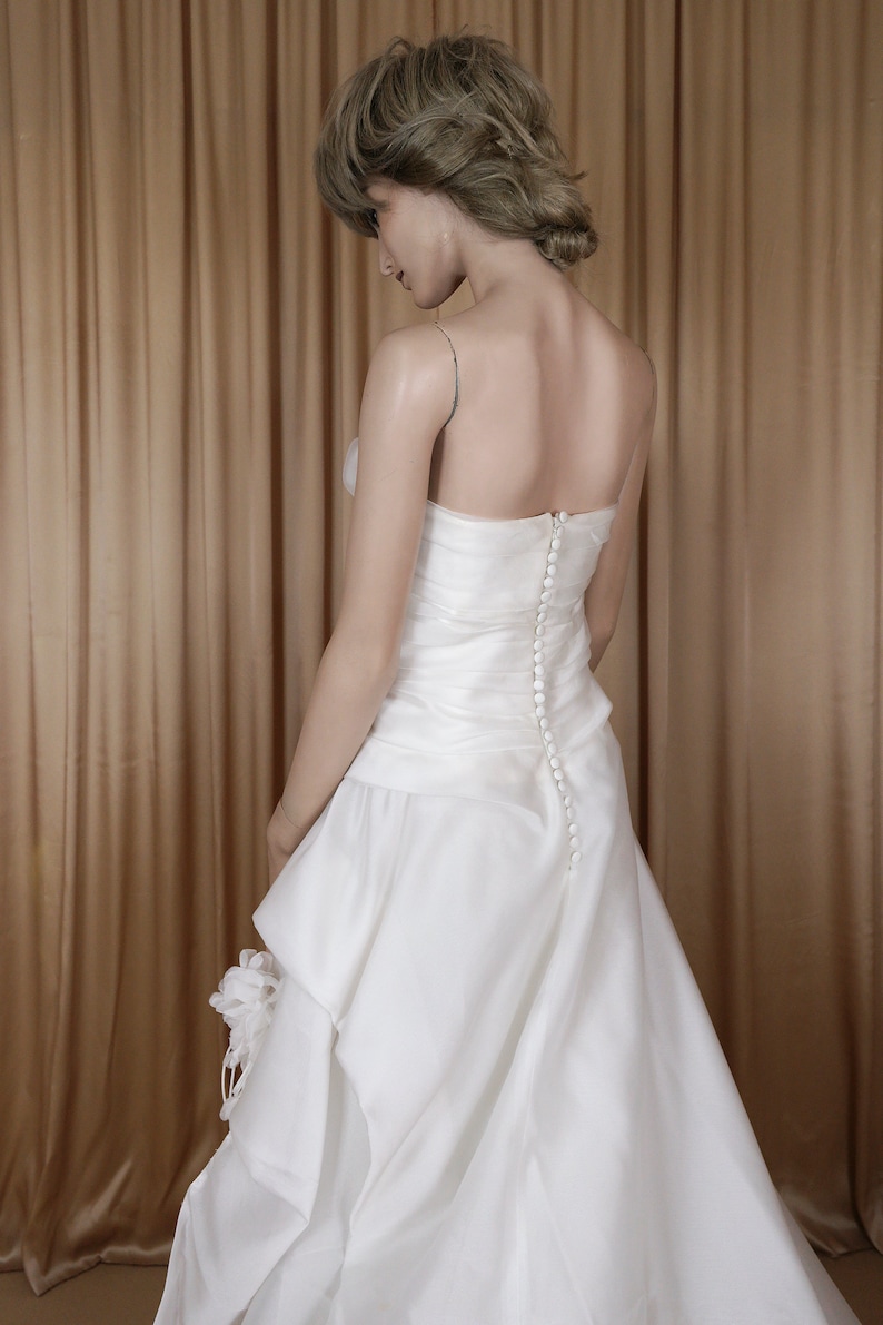 May include: A white bridal gown with a fitted bodice and a flowing skirt. The back of the dress has a row of small buttons. The dress features thin straps and a layered skirt detail. The mannequin is positioned in front of a gold curtain.