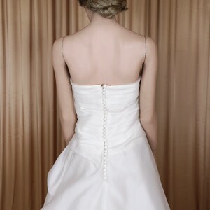 May include: A white, strapless wedding dress with a fitted bodice and a flowing skirt. The back of the dress has a row of small, round buttons. Delicate silver straps are visible.