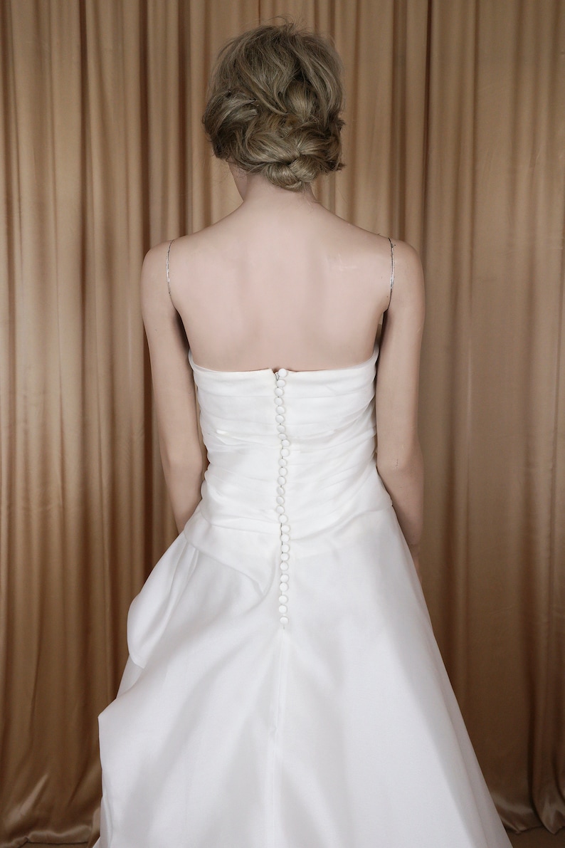 May include: A white, strapless wedding dress with a fitted bodice and a flowing skirt. The back of the dress has a row of small, round buttons. Delicate silver straps are visible.