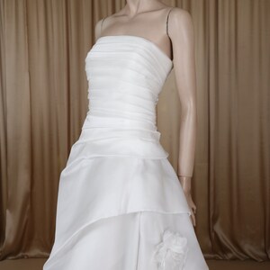 May include: A white strapless wedding dress on a mannequin. The bodice has horizontal pleats, and the skirt features layered fabric with a floral accent. The dress has a flowing train, ideal for a bridal look.