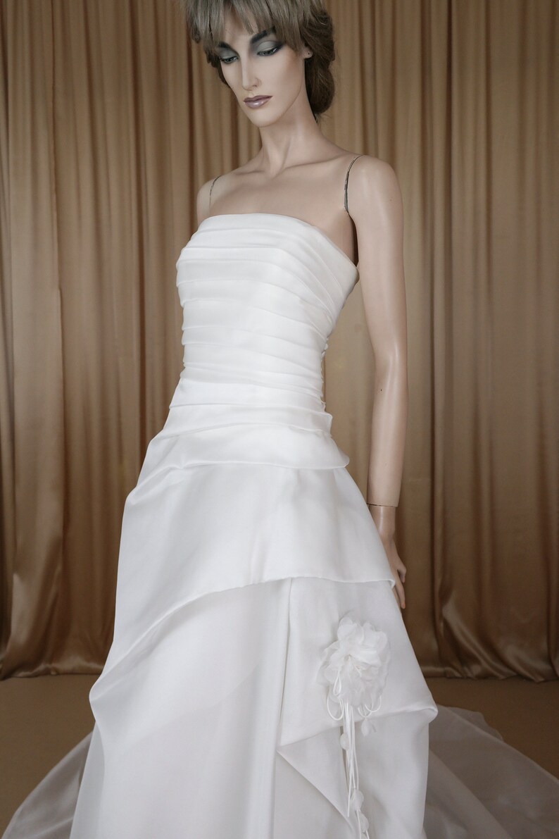 May include: A white strapless wedding dress on a mannequin. The bodice has horizontal pleats, and the skirt features layered fabric with a floral accent. The dress has a flowing train, ideal for a bridal look.
