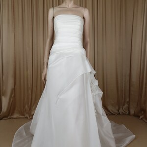 May include: A white strapless wedding dress on a mannequin. The bodice has horizontal pleats, and the skirt features layered fabric with a flowing train. The dress is displayed against a neutral background.