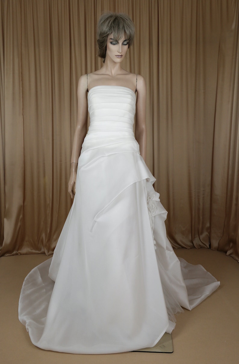 May include: A white strapless wedding dress on a mannequin. The bodice has horizontal pleats, and the skirt features layered fabric with a flowing train. The dress is displayed against a neutral background.