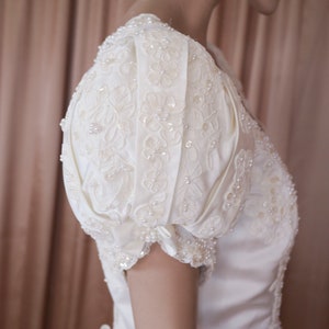 May include: A white satin wedding dress with a fitted bodice and a full skirt. The dress features a delicate lace overlay with pearl embellishments and a puff sleeve design.