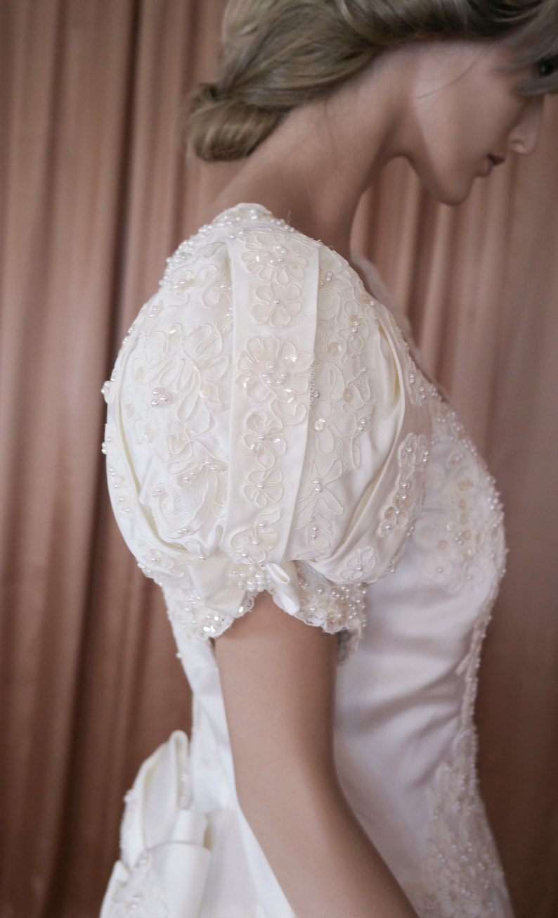 May include: A white satin wedding dress with a fitted bodice and a full skirt. The dress features a delicate lace overlay with pearl embellishments and a puff sleeve design.