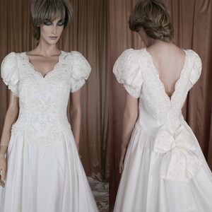 May include: A white satin wedding dress with a lace bodice and a large bow at the back. The dress has puffed sleeves and a v-neckline.