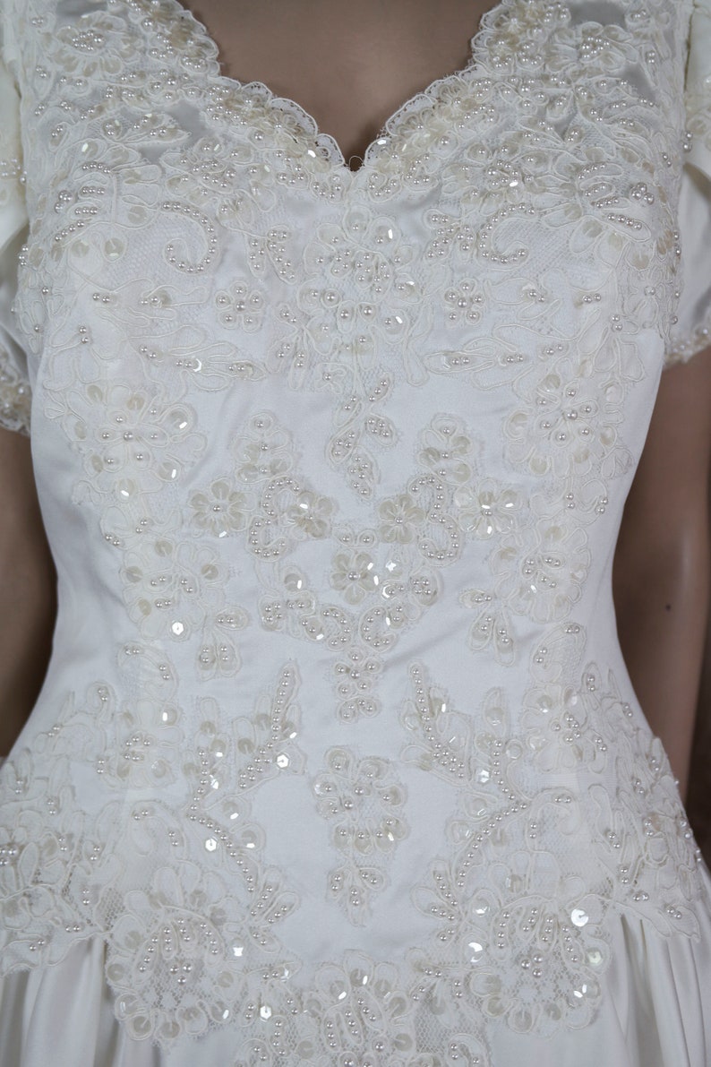 May include: A white satin wedding dress with a lace bodice and pearl embellishments. The dress has a sweetheart neckline and short sleeves.