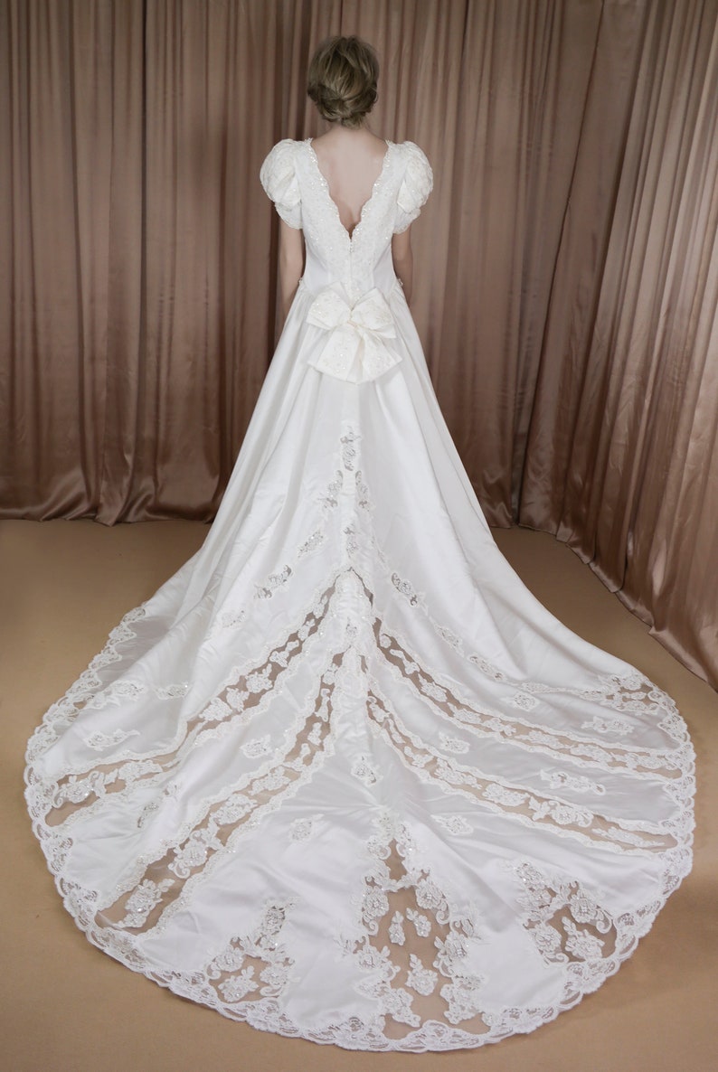 May include: A white satin wedding dress with a lace overlay and a large bow at the back.