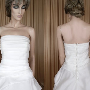 May include: A white strapless wedding dress with a layered bodice and a flowing skirt. The dress has a row of buttons down the back. The fabric appears to be satin or a similar material. The dress is displayed on a mannequin.
