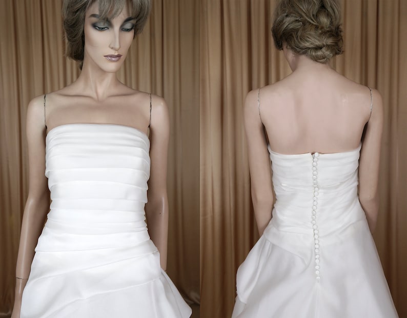 May include: A white strapless wedding dress with a layered bodice and a flowing skirt. The dress has a row of buttons down the back. The fabric appears to be satin or a similar material. The dress is displayed on a mannequin.