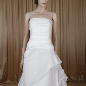 May include: A white strapless wedding dress on a mannequin. The bodice has horizontal pleats, and the skirt features layered fabric with a floral accent. The dress is floor-length, with a flowing train. The mannequin has a neutral expression.