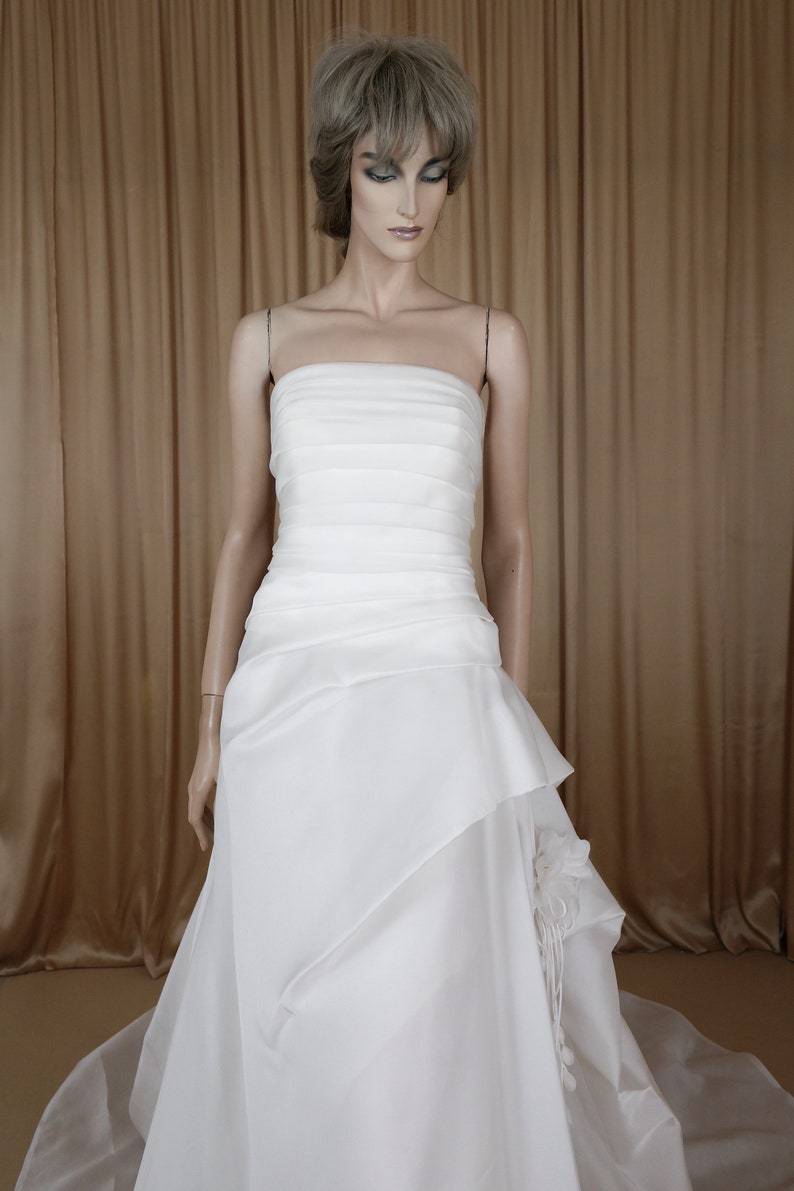 May include: A white strapless wedding dress on a mannequin. The bodice has horizontal pleats, and the skirt features layered fabric with a floral accent. The dress is floor-length, with a flowing train. The mannequin has a neutral expression.