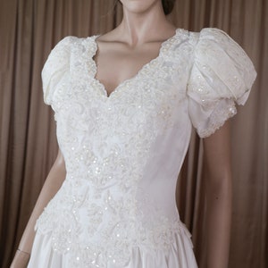 May include: A white satin wedding dress with a sweetheart neckline and short puff sleeves. The bodice is adorned with intricate beading and lace.
