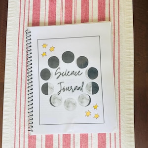 May include: A spiral-bound science journal with a white cover featuring a black and white illustration of the phases of the moon. The words "Science Journal" are written in cursive. Yellow stars are scattered around the moon phases.