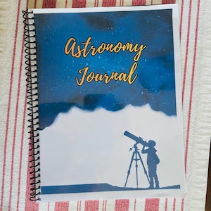 May include: A spiral-bound journal with a cover featuring a dark blue night sky with stars and the text Astronomy Journal in yellow script. The lower portion of the cover is white and displays a silhouette of a person using a telescope.