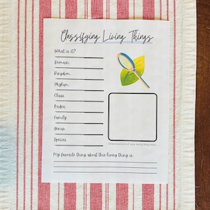 Puede incluir: Una hoja de papel blanca con el título "Classifying Living Things" en escritura azul. El papel incluye líneas para completar la clasificación científica, un cuadro de dibujo y una ilustración de lupa.