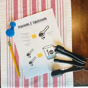 以下が含まれることがあります： 「TEASPOONS & TABLESPOONS」の文字と、計量スプーンと計量カップのイラストが描かれた白い紙。青いマグネット、黄色の鉛筆、黒い計量スプーンのセットも表示されています。