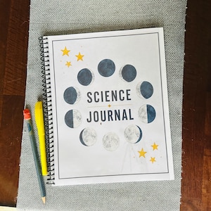May include: A spiral-bound science journal with a white cover featuring a circular arrangement of moon phases in shades of blue and gray, the text "SCIENCE JOURNAL" in dark blue, and small yellow stars, accompanied by a yellow pen and a gray pencil.