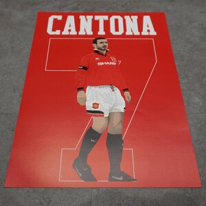 May include: A red poster featuring a stylized illustration of a soccer player in a red jersey with the name "CANTONA" above. The player wears white shorts and black socks, with the number "7" prominently displayed.