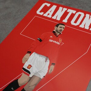 May include: Red poster featuring a stylized illustration of a soccer player in a red jersey with the name "CANTONA" in white block letters. The player is wearing white shorts and black socks. The jersey has the "SHARP" logo.