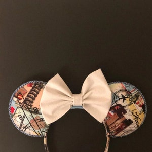 May include: A pair of white fabric mouse ears with a blue and white patterned fabric bow. The ears are decorated with a travel theme and feature images of airplanes, maps, and luggage.