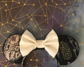 May include: A pair of black mouse ears with a white bow. One ear features a black and white image of the Millennium Falcon from Star Wars. The other ear features the text "Star Wars" in yellow.