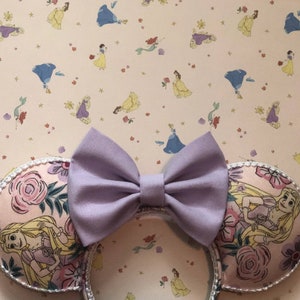 May include: A pair of purple fabric Minnie Mouse ears with a floral print featuring Rapunzel. The ears are decorated with a large purple bow and have a white background with a floral pattern.