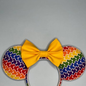May include: Rainbow Mickey Mouse ears headband with a yellow bow. The headband is made of fabric with a rainbow pattern of Mickey Mouse heads.