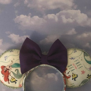 May include: Purple bow Minnie Mouse ears with a white background featuring a mermaid, bubbles, and the text "Free as the Sea".