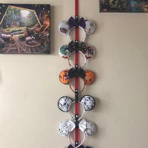 May include: A red ribbon hanging on a wall with seven pairs of colorful mouse ears hanging from it. The mouse ears are decorated with various Disney characters and designs.