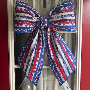May include: A large quilted fabric bow featuring a red, white, and blue color scheme with repeating patterns of the word Bills and buffalo logos. The bow is constructed from multiple strips of fabric with visible stitching and hangs from a brown strap.