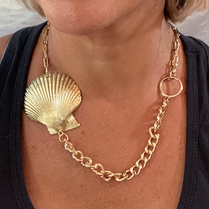 May include: A gold chain necklace with a large, gold seashell pendant. The chain has a large, round, gold ring at the top.