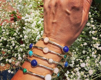May include: A collection of gold-toned bangles, each adorned with a different gemstone. The gemstones include blue lapis lazuli, turquoise, malachite, and rose quartz. The bangles are displayed against a backdrop of white flowers.