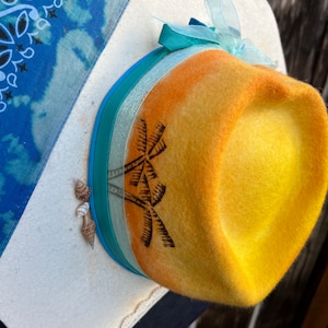 May include: A yellow fedora-style hat with an orange gradient, featuring a palm tree design. The hat is adorned with blue and white ribbons and a sheer turquoise bow. Small seashells are attached to the brim, evoking a beach theme.