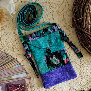 May include: A small teal and purple patchwork crossbody bag with a floral print and a black button. The bag has a long strap and a small pocket on the front. A black circle with a gold butterfly is embroidered on the front of the bag.