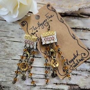 May include: Handmade earrings with woven fabric tops in gold and white, accented with brown and pink threads. The earrings feature dangling chains with amber-colored beads and brass-toned buttons. The tag reads "The Hypno Fae Recycled Jewelry."