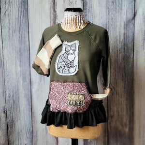 May include: A green sweatshirt with a black and white fox graphic on the front. The sweatshirt has a burgundy and black patchwork design on the bottom and a black ruffle trim.