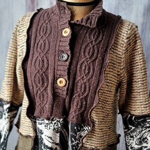 May include: A brown and tan patchwork sweater with a cable-knit collar and button closure. The sweater features a mix of textures and patterns, including a patterned sleeve and a decorative trim.