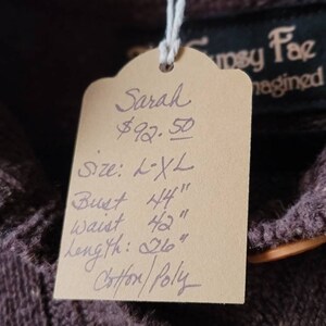 May include: A brown garment with a tag that reads "Sarah" and lists measurements: Bust 44", Waist 42", Length 26". The tag also indicates the size is L-XL and the material is Cotton/Poly. The brand name "Gyspy Fae" is visible.