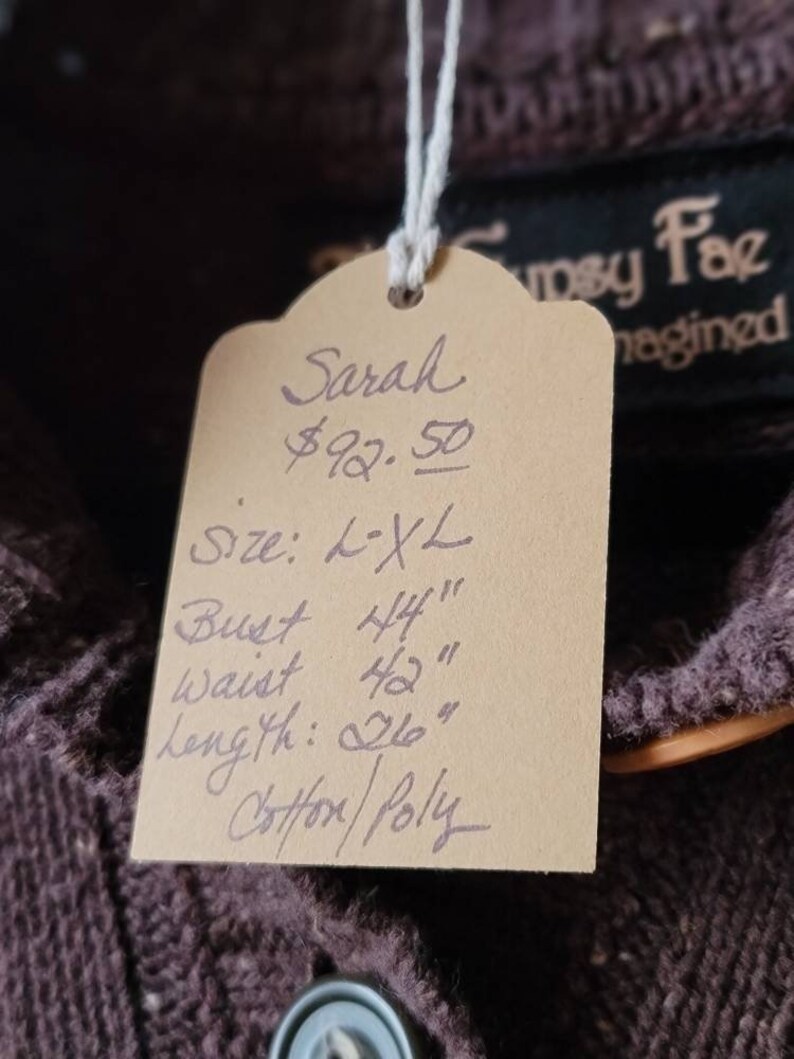 May include: A brown garment with a tag that reads "Sarah" and lists measurements: Bust 44", Waist 42", Length 26". The tag also indicates the size is L-XL and the material is Cotton/Poly. The brand name "Gyspy Fae" is visible.