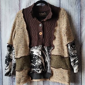 May include: A patchwork sweater with a brown, tan, and black color scheme. The sweater features a cable knit collar and button closure. The sleeves have a black and white patterned fabric. The text "The Gypsy Fox Clothing Remade" is visible on the collar.
