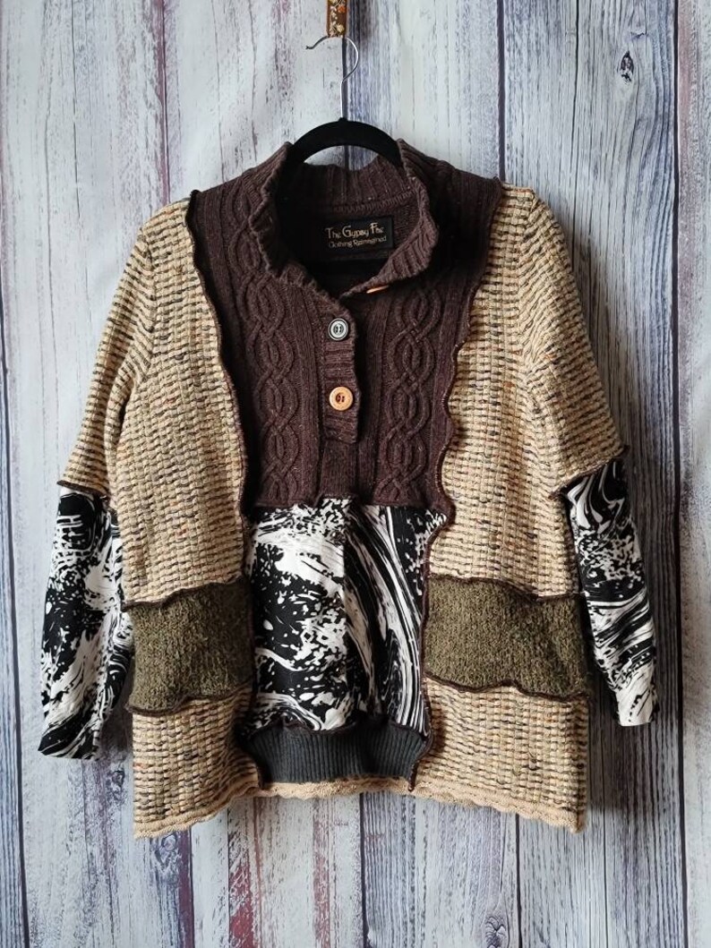 May include: A patchwork sweater with a brown, tan, and black color scheme. The sweater features a cable knit collar and button closure. The sleeves have a black and white patterned fabric. The text "The Gypsy Fox Clothing Remade" is visible on the collar.