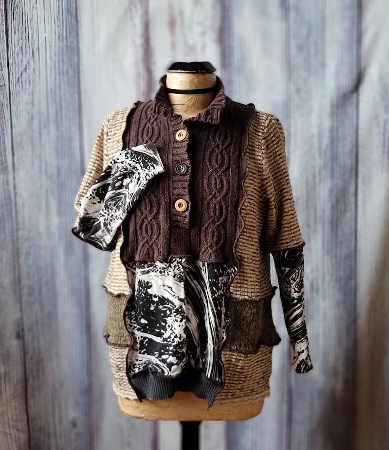 May include: A unique patchwork sweater displayed on a mannequin. The sweater features a brown cable-knit front with wooden buttons, layered with patterned fabrics in black, white, and olive green. The sleeves have a black and white swirl pattern.