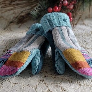 May include: A pair of knit mittens featuring a patchwork design with panels of light blue, gray, mustard yellow, pink, and a dark blue patterned trim. The mittens have a ribbed light blue cuff with a small matching button on the outer edge.