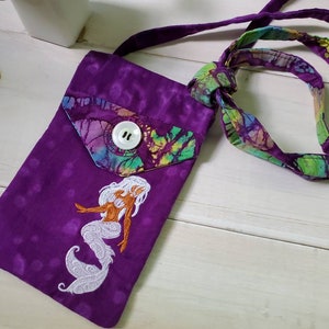 May include: A purple fabric crossbody bag with a mermaid embroidery. The bag has a flap with a button closure and a colorful batik pattern. The strap is also made of batik fabric. The mermaid has white hair and a white tail.