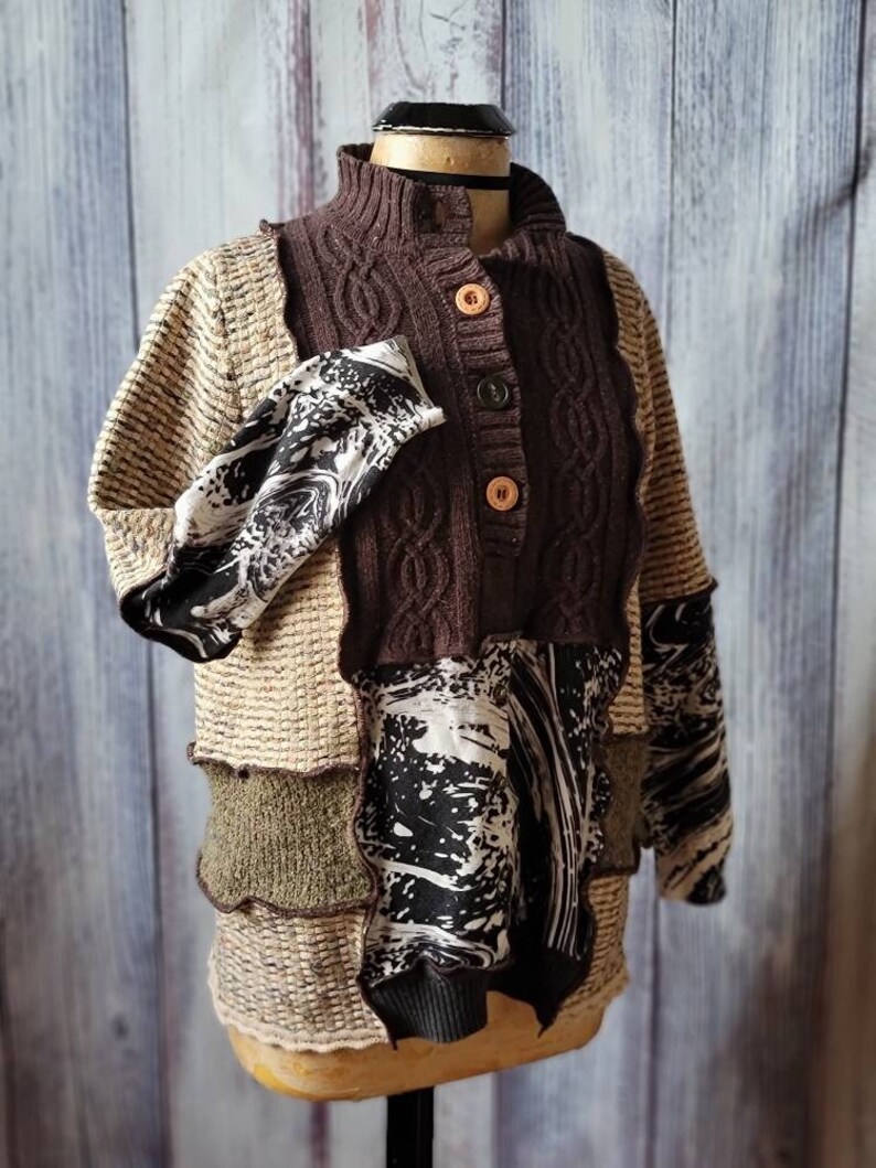 May include: A patchwork sweater displayed on a mannequin. The sweater features a brown cable-knit bodice with wooden buttons, tan sleeves, and panels of olive green and black and white patterned fabric. The sweater has a high collar.