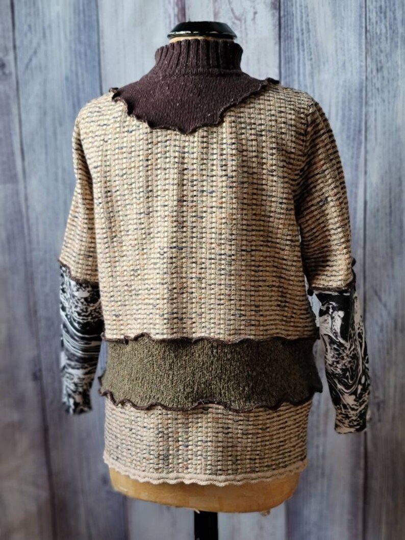 May include: A textured sweater with a brown turtleneck and sleeves with a black and white pattern. The body of the sweater is beige with brown and black flecks, and a green band across the midsection. The bottom edge has a scalloped trim.