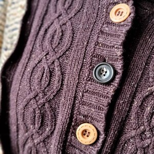 May include: Close-up of a dark purple cable-knit cardigan with three buttons. The buttons are a light tan color, and the cardigan has a textured, woven appearance. The cardigan is a warm, cozy garment.