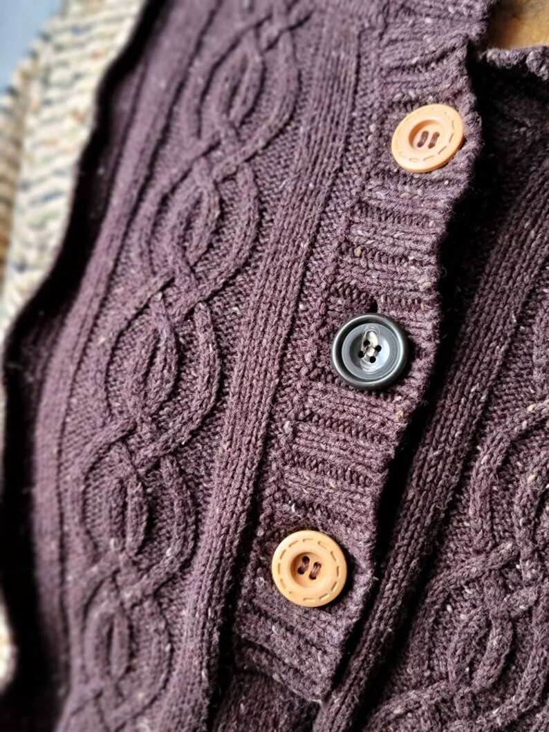 May include: Close-up of a dark purple cable-knit cardigan with three buttons. The buttons are a light tan color, and the cardigan has a textured, woven appearance. The cardigan is a warm, cozy garment.