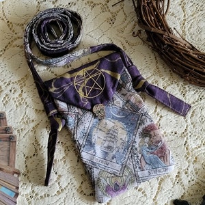 May include: A purple tarot card bag with a gold embroidered pentagram and a skull button. The bag features tarot card illustrations and a long strap. The bag is made of fabric and is designed to hold tarot cards.